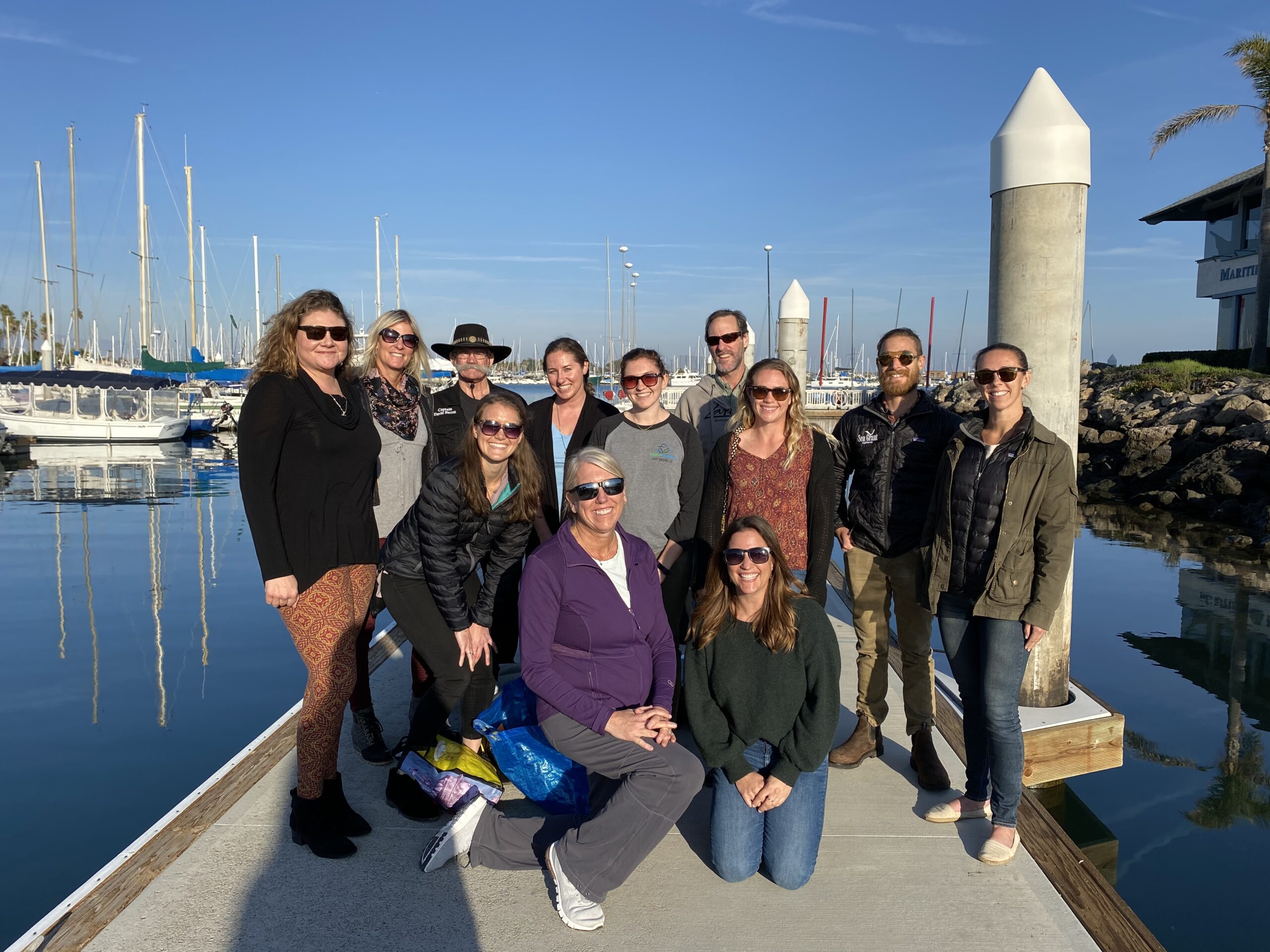 Photo shows a group of MPA CN members posing together standing on a dock with ocean harbor in the background/