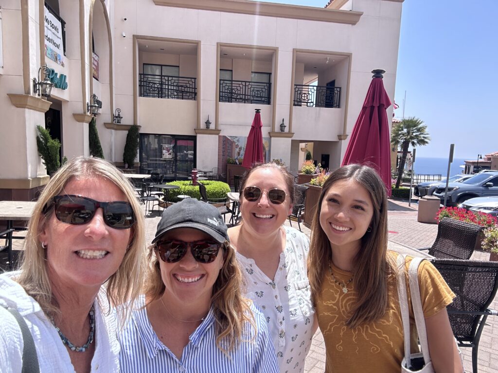 Left to right: Calla Allison, Aubrie Fowler, Claire Arre, and Lea Graham at a LA. MPA Collaborative Meeting smiling at the camera in a courtyard with the ocean in the background.